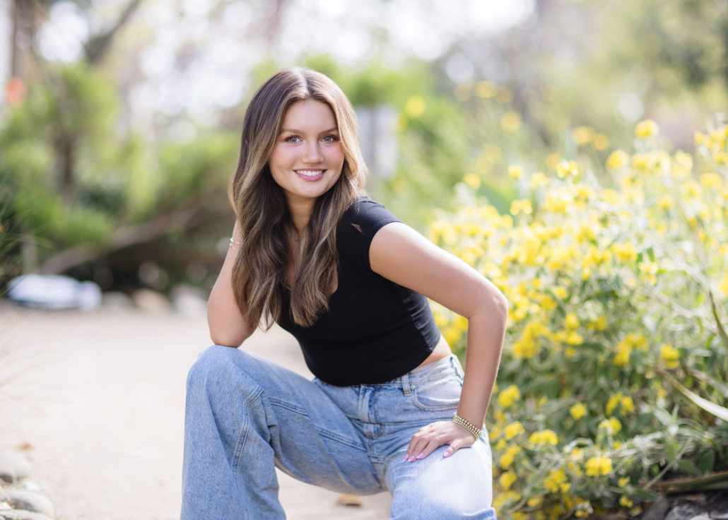Kyra Walsh sitting on the ground in a casual outfit of boyfriend jeans and a black crop top, with a vibrant outdoor setting in San Juan Capistrano, showcasing her personality in her senior photoshoot