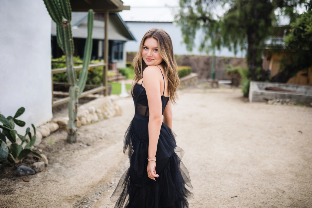 Kyra Walsh in an elegant black dress, looking over her shoulder and smiling, standing on a pathway surrounded by historic buildings and cactus plants in San Juan Capistrano, showcasing a blend of modern style and historical charm during her senior photoshoot