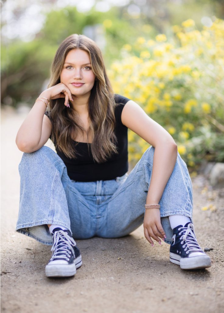 Kyra Walsh sitting on the ground in a casual outfit of boyfriend jeans and a black crop top, with a vibrant outdoor setting in San Juan Capistrano, showcasing her personality in her senior photoshoot