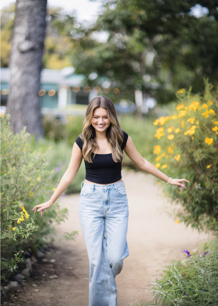 Kyra Walsh walking along a garden path in San Juan Capistrano, wearing a black crop top and high-waisted jeans, smiling and surrounded by vibrant yellow flowers and greenery, capturing a candid moment during her senior photoshoot