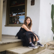 Kyra Walsh sitting on the steps of a rustic building, wearing a black dress with Converse sneakers, during her senior photography session in San Juan Capistrano