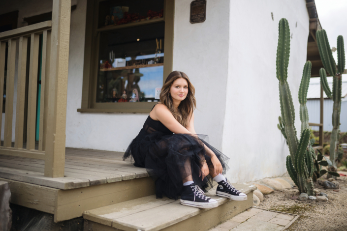 Kyra Walsh sitting on the steps of a rustic building, wearing a black dress with Converse sneakers, during her senior photography session in San Juan Capistrano