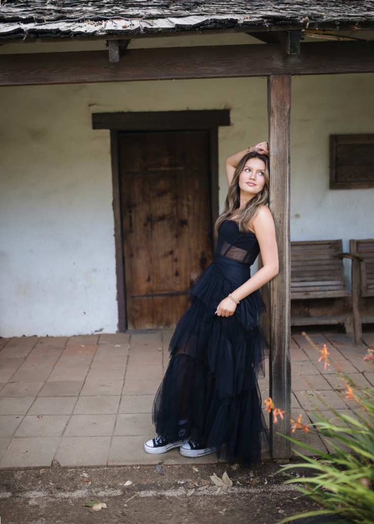 Kyra Walsh leaning against a rustic wooden building in Historic San Juan Capistrano, wearing an elegant black dress, highlighting her senior photoshoot style