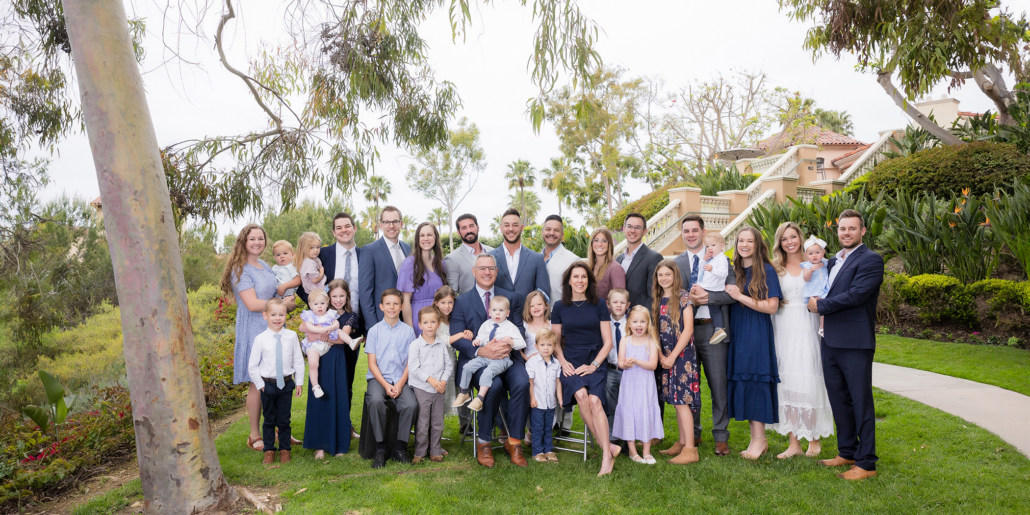 A large family portrait taken outdoors in Laguna Beach. The group consists of several families, including men, women, and children of various ages, dressed in formal attire. They are standing and sitting on a lush green lawn, with a backdrop of trees, bushes, and a beautiful house with a terracotta roof. The group is smiling and posing together under an overcast sky.