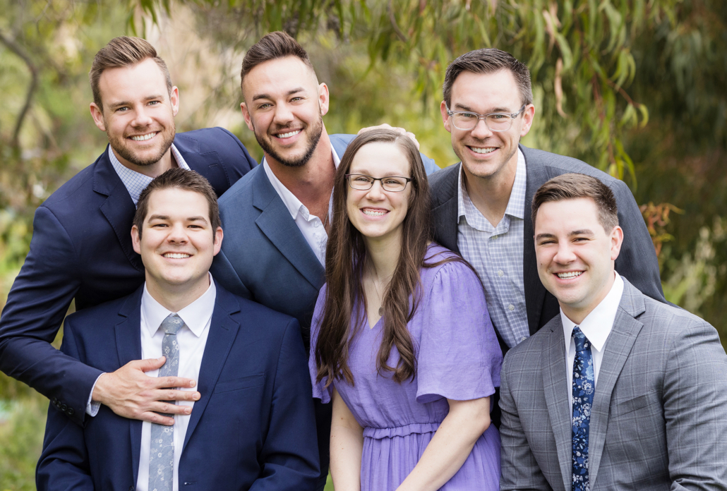 A group portrait of six adult siblings taken at the Newport Coast Marriott. They are standing closely together outdoors, smiling warmly at the camera. The siblings are dressed in formal attire: three men in navy blue suits, one man in a gray suit with a blue floral tie, one man in a gray blazer with a checked shirt, and a woman in a light purple dress. The background features lush greenery, adding a natural and serene ambiance to the portrait. This image captures a moment of family bonding and joy