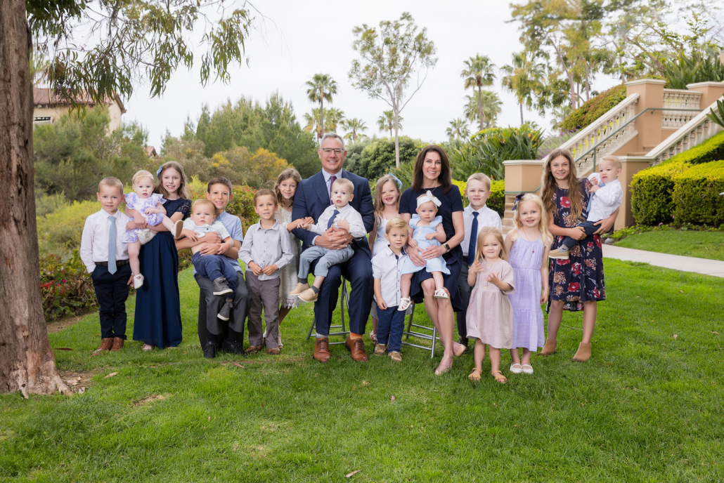 A large extended family portrait taken in Newport Coast, featuring grandparents with their numerous grandchildren. The grandparents are seated in the center, holding some of the younger grandchildren. The group is dressed in semi-formal attire, with the children wearing a mix of dresses, suits, and casual outfits. The lush greenery, palm trees, and elegant staircase in the background add to the scenic outdoor setting. The family is gathered on a well-manicured lawn, capturing a moment of joy and togetherness with the grandparents surrounded by their loving grandchildren.
