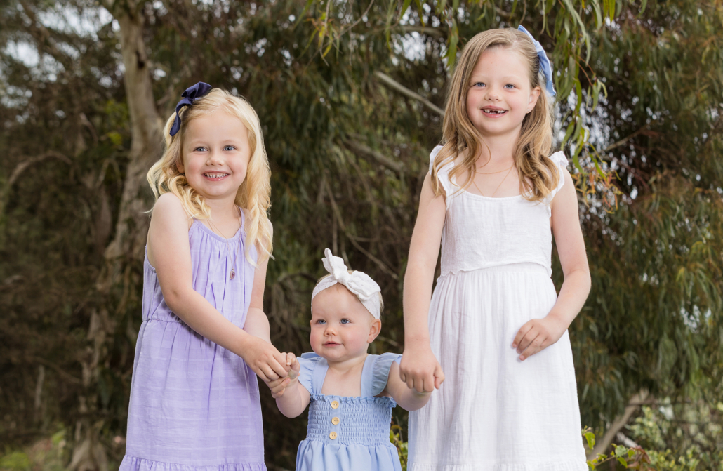 A portrait of three young sisters taken outdoors in Newport Coast. The eldest sister, wearing a white dress with a light blue bow in her hair, and the middle sister, in a light purple dress with a navy bow, are holding hands with their baby sister. The baby sister is dressed in a light blue dress with a white headband. The girls are smiling and standing together in front of a backdrop of lush greenery, capturing a moment of sibling affection and joy. This image highlights the close bond and happiness of the sisters in a natural setting.