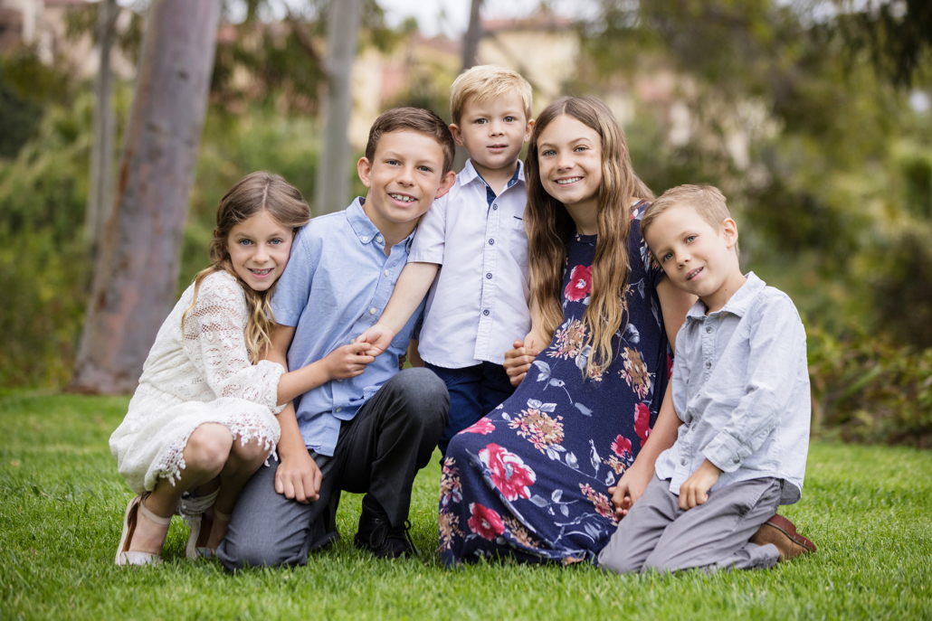A portrait of five young children taken outdoors in Newport Coast. The children are sitting and kneeling on a lush green lawn, smiling and looking at the camera. The group includes two girls and three boys. The girls are dressed in a white lace dress and a navy blue floral dress, while the boys are wearing light blue and white shirts with gray pants. The background features trees and a blurred view of buildings, creating a serene and natural setting. This image captures the children's happiness and close bond