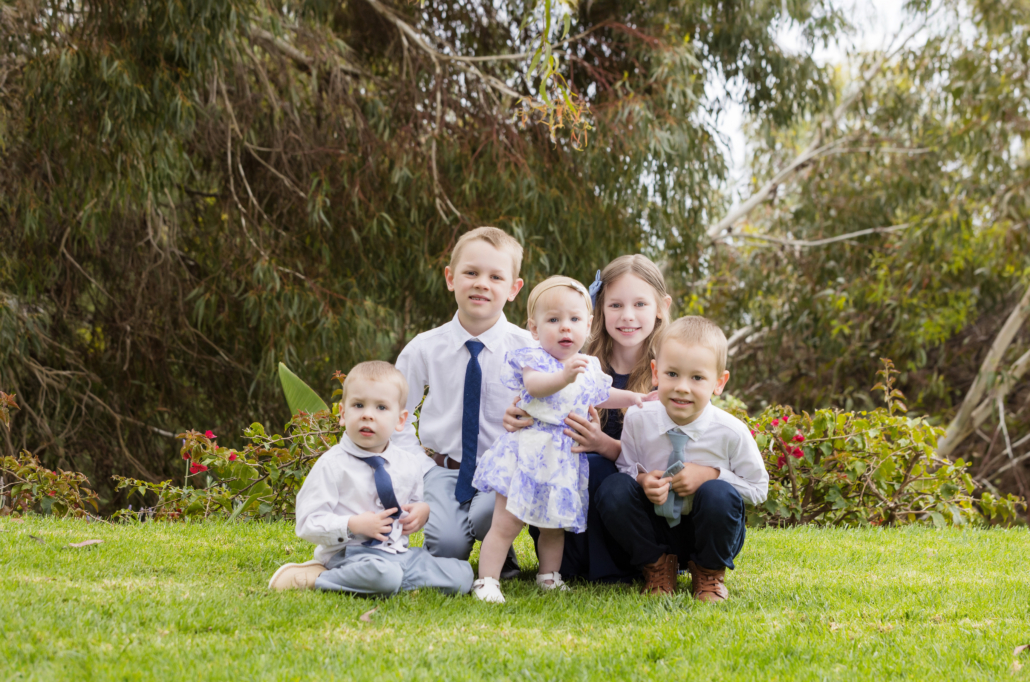  portrait of five young siblings taken outdoors in Orange County. The children are sitting and kneeling on a lush green lawn, smiling and looking at the camera. The group includes two boys dressed in white shirts and ties, a girl in a navy blue dress with a light blue bow, a toddler boy in a white shirt with a navy tie, and a baby girl in a white and blue floral dress with a headband. The background features lush greenery and flowering plants, creating a natural and serene setting. This image captures the siblings' close bond and joyful expressions.