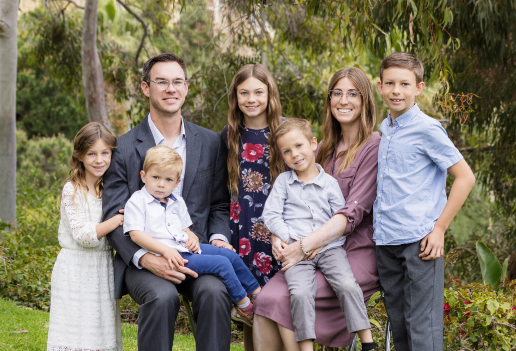 A portrait of a large Mormon family taken in Newport Coast, outdoors, featuring a mother, father, and their five children. The family is dressed in semi-formal attire: the father in a gray suit, the mother in a mauve dress, and the children in various outfits including a white lace dress, a floral dress, and light-colored shirts and pants. The family members are sitting and standing close together on a lush green lawn with trees and shrubs in the background. They are smiling and looking at the camera, capturing a moment of familial love and togetherness in a natural setting.