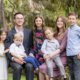 A portrait of a large Mormon family taken in Newport Coast, outdoors, featuring a mother, father, and their five children. The family is dressed in semi-formal attire: the father in a gray suit, the mother in a mauve dress, and the children in various outfits including a white lace dress, a floral dress, and light-colored shirts and pants. The family members are sitting and standing close together on a lush green lawn with trees and shrubs in the background. They are smiling and looking at the camera, capturing a moment of familial love and togetherness in a natural setting.