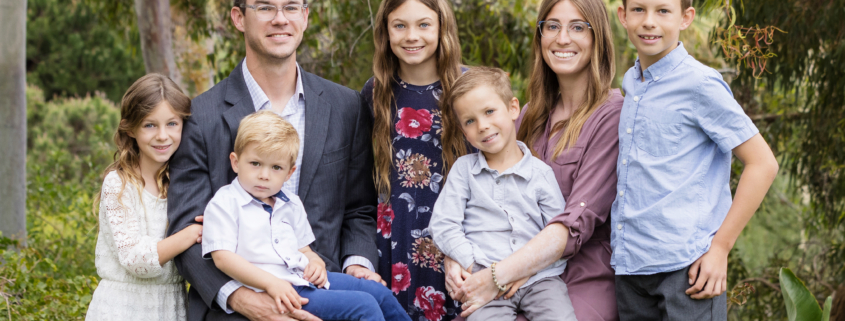 A portrait of a large Mormon family taken in Newport Coast, outdoors, featuring a mother, father, and their five children. The family is dressed in semi-formal attire: the father in a gray suit, the mother in a mauve dress, and the children in various outfits including a white lace dress, a floral dress, and light-colored shirts and pants. The family members are sitting and standing close together on a lush green lawn with trees and shrubs in the background. They are smiling and looking at the camera, capturing a moment of familial love and togetherness in a natural setting.