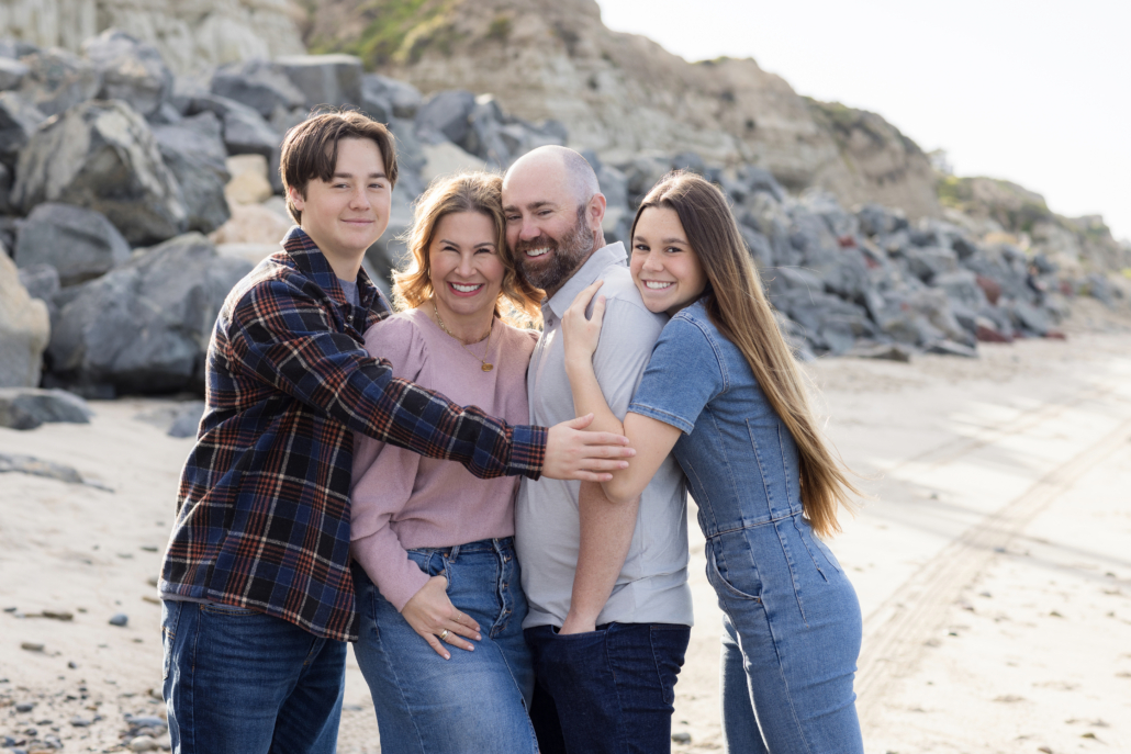 Family of four embracing in a loving hug with the scenic San Clemente Beach behind them.