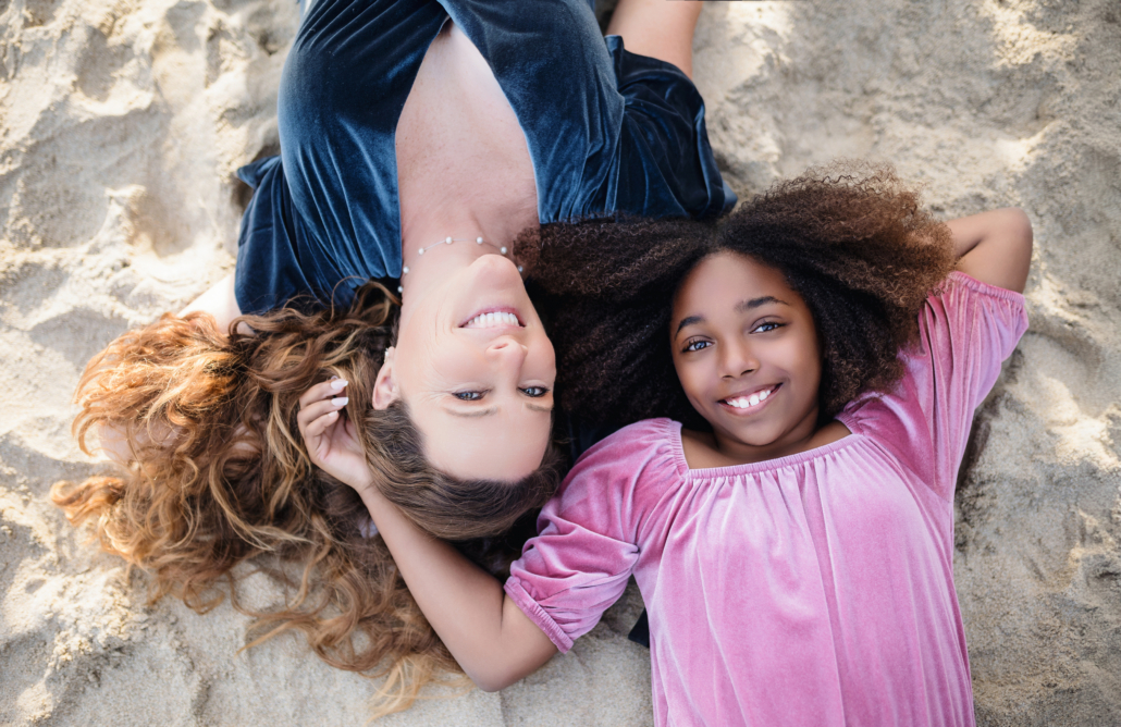 Mother and daughter lying on the beach, heads close together, smiling up at the sky as the sun casts a warm glow on them.