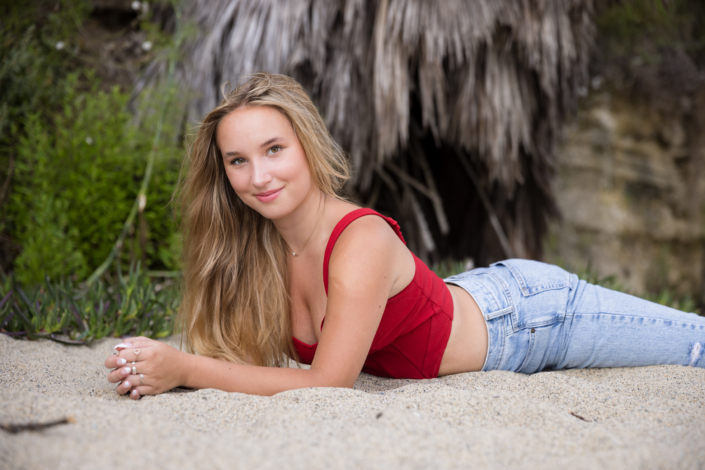 A high school senior girl with long blonde hair poses on the sandy beach, leaning on her elbows and smiling softly. She wears a stylish red crop top and light-wash jeans, with a natural, beachy backdrop of palm fronds and greenery behind her.