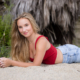 A high school senior girl with long blonde hair poses on the sandy beach, leaning on her elbows and smiling softly. She wears a stylish red crop top and light-wash jeans, with a natural, beachy backdrop of palm fronds and greenery behind her.