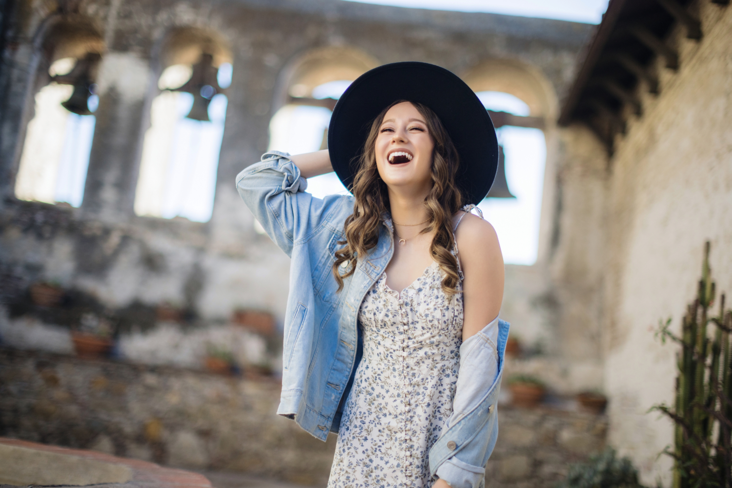 A high school senior girl laughs joyfully while posing at a historic mission. She wears a black wide-brimmed hat, a floral dress, and a light denim jacket draped over her shoulders. The weathered stone arches and hanging bells in the background add a timeless charm to the scene. Mission San Juan. Relaxed poses 