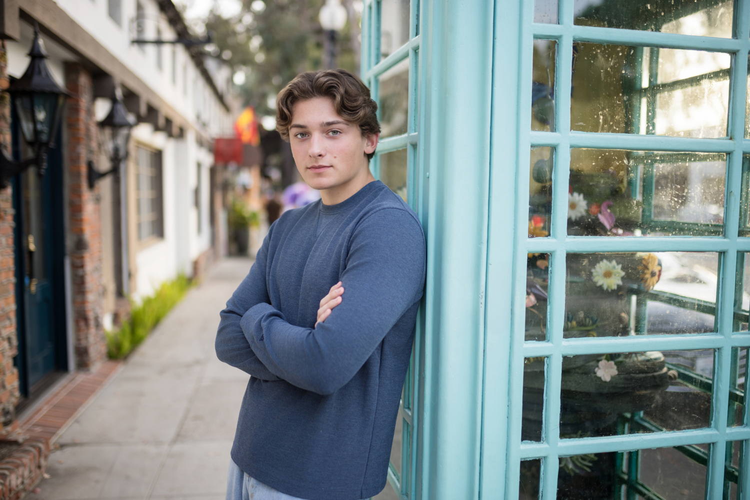 A high school senior guy leans against a vintage turquoise phone booth in downtown Laguna Beach, arms crossed with a confident expression. Dressed in a casual blue sweater and light-wash jeans, he stands against a charming backdrop of brick storefronts and European-style architecture, creating a stylish and timeless urban senior portrait.