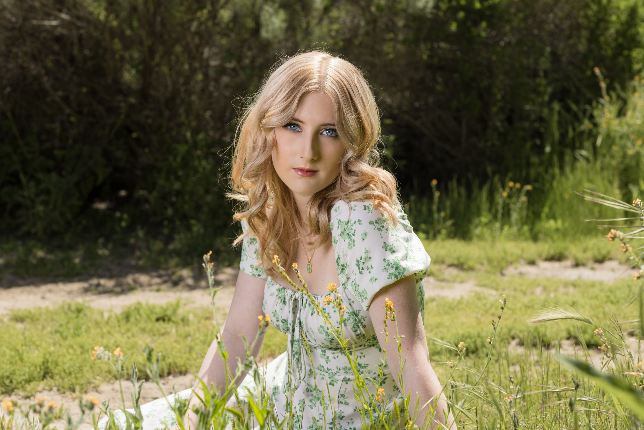 High school senior girl with blonde curls wearing a white and green floral dress, sitting in a field of wildflowers with a soft, natural backdrop. A dreamy outdoor senior portrait in Orange County.