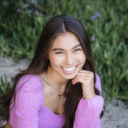 High school senior smiling brightly in a Laguna Beach outdoor portrait session. Wearing a vibrant pink sweater, she poses naturally with lush greenery in the background, captured by an Orange County senior photographer.