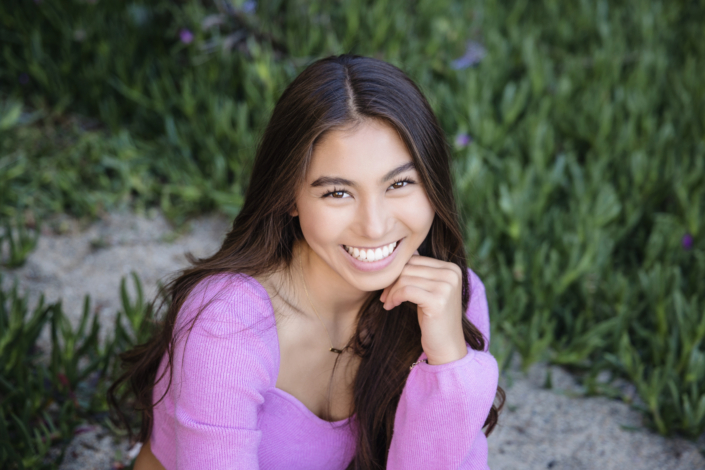 High school senior smiling brightly in a Laguna Beach outdoor portrait session. Wearing a vibrant pink sweater, she poses naturally with lush greenery in the background, captured by an Orange County senior photographer.