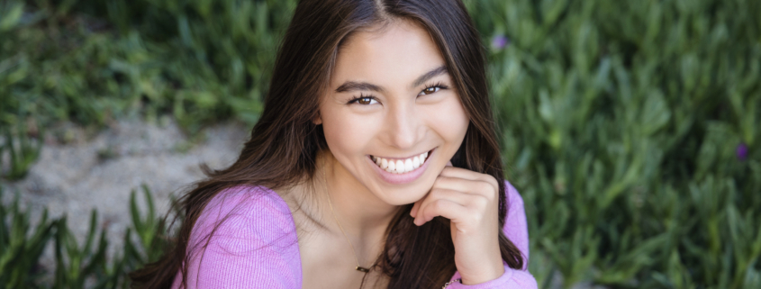 High school senior smiling brightly in a Laguna Beach outdoor portrait session. Wearing a vibrant pink sweater, she poses naturally with lush greenery in the background, captured by an Orange County senior photographer.