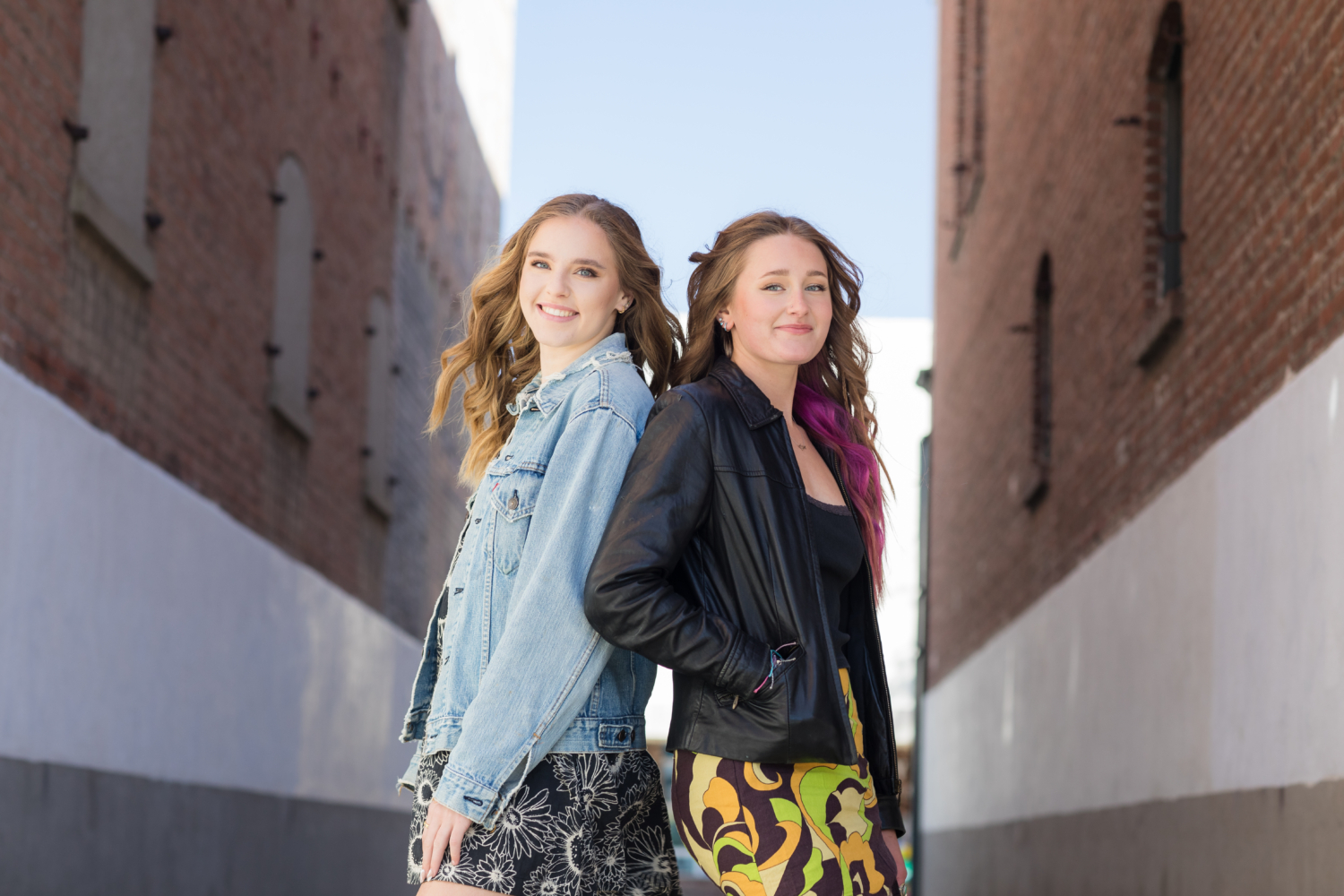 Two stylish high school seniors standing back-to-back in an urban alleyway, smiling confidently. One wears a denim jacket and floral skirt, while the other sports a black leather jacket and a bold patterned skirt, showcasing a mix of casual and edgy styles.