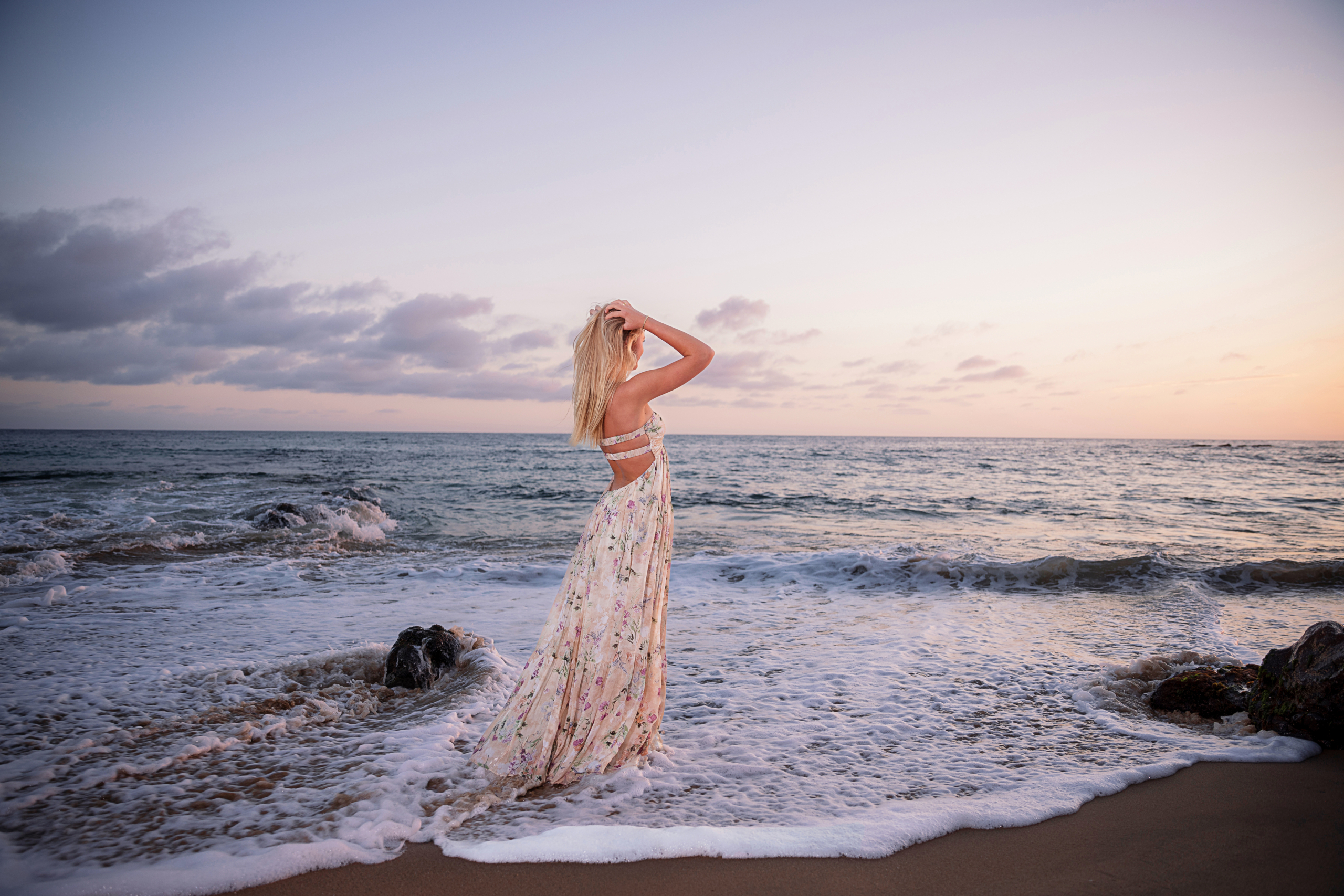 A young woman in a flowing floral dress stands in the ocean waves at sunset, gazing toward the horizon with soft golden light illuminating her hair.