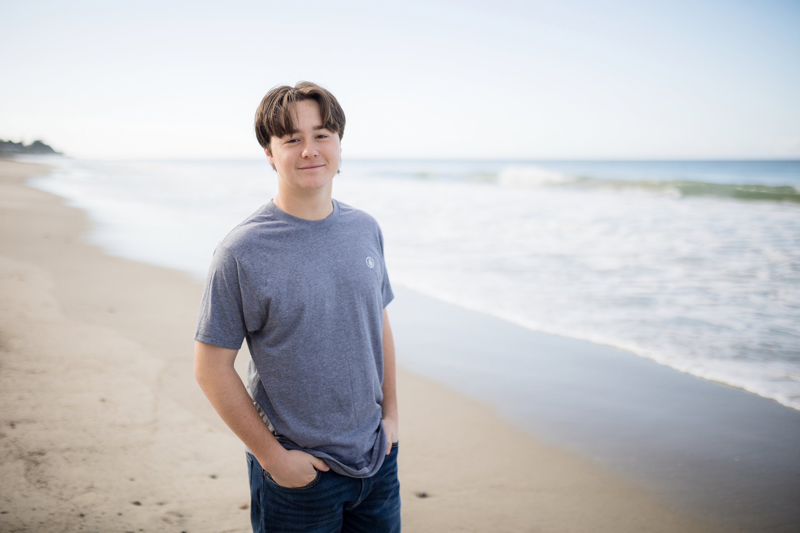 Senior boy standing on the beach in a casual gray t-shirt and jeans, hands in pockets, smiling softly with ocean waves gently rolling in behind him under a bright, clear sky.