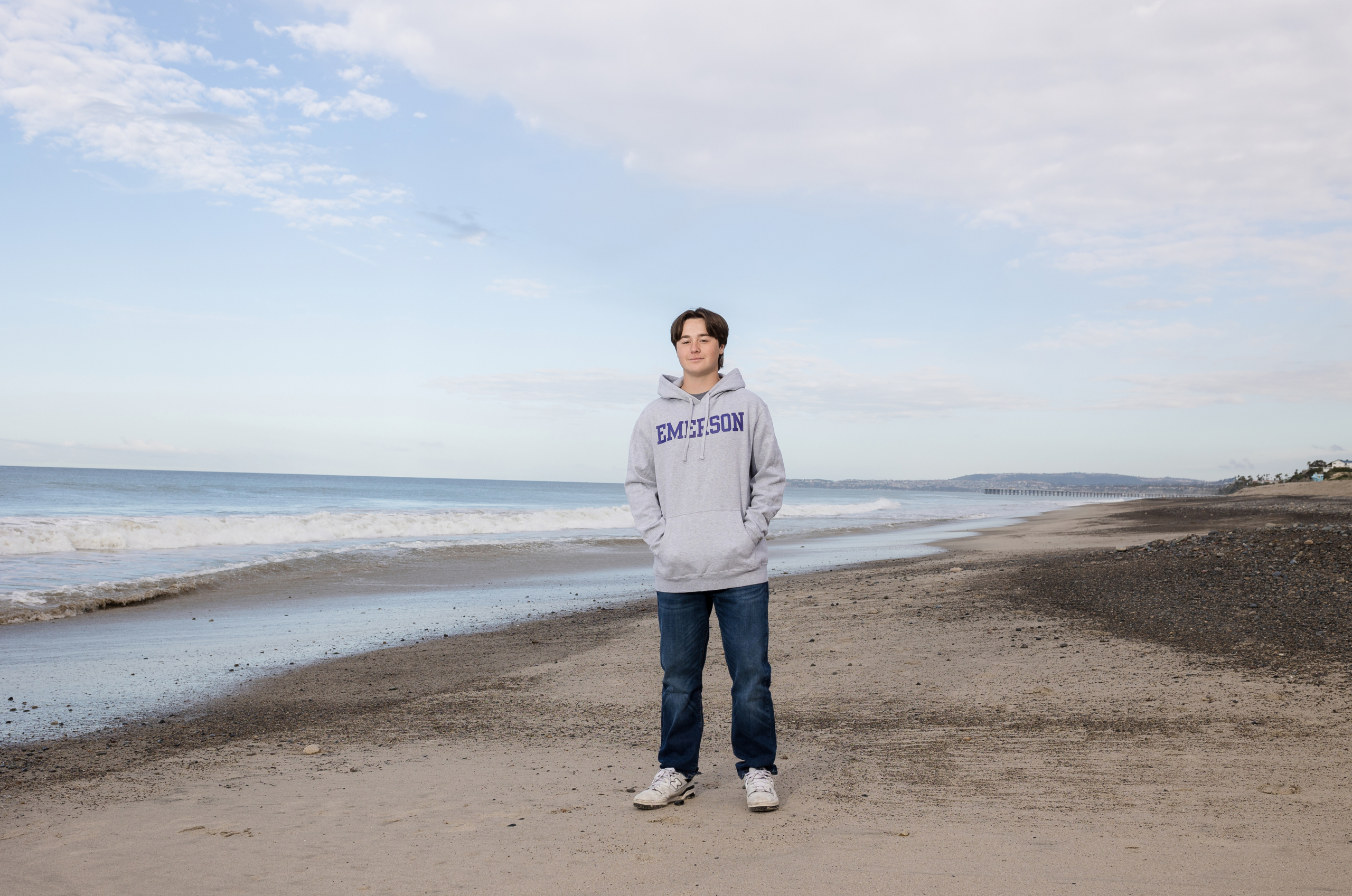 A high school senior leans casually against a lifeguard tower on the sand, dressed in a black jacket over a white button-up shirt. The ocean, cliffs, and beachfront homes add depth to the coastal setting.