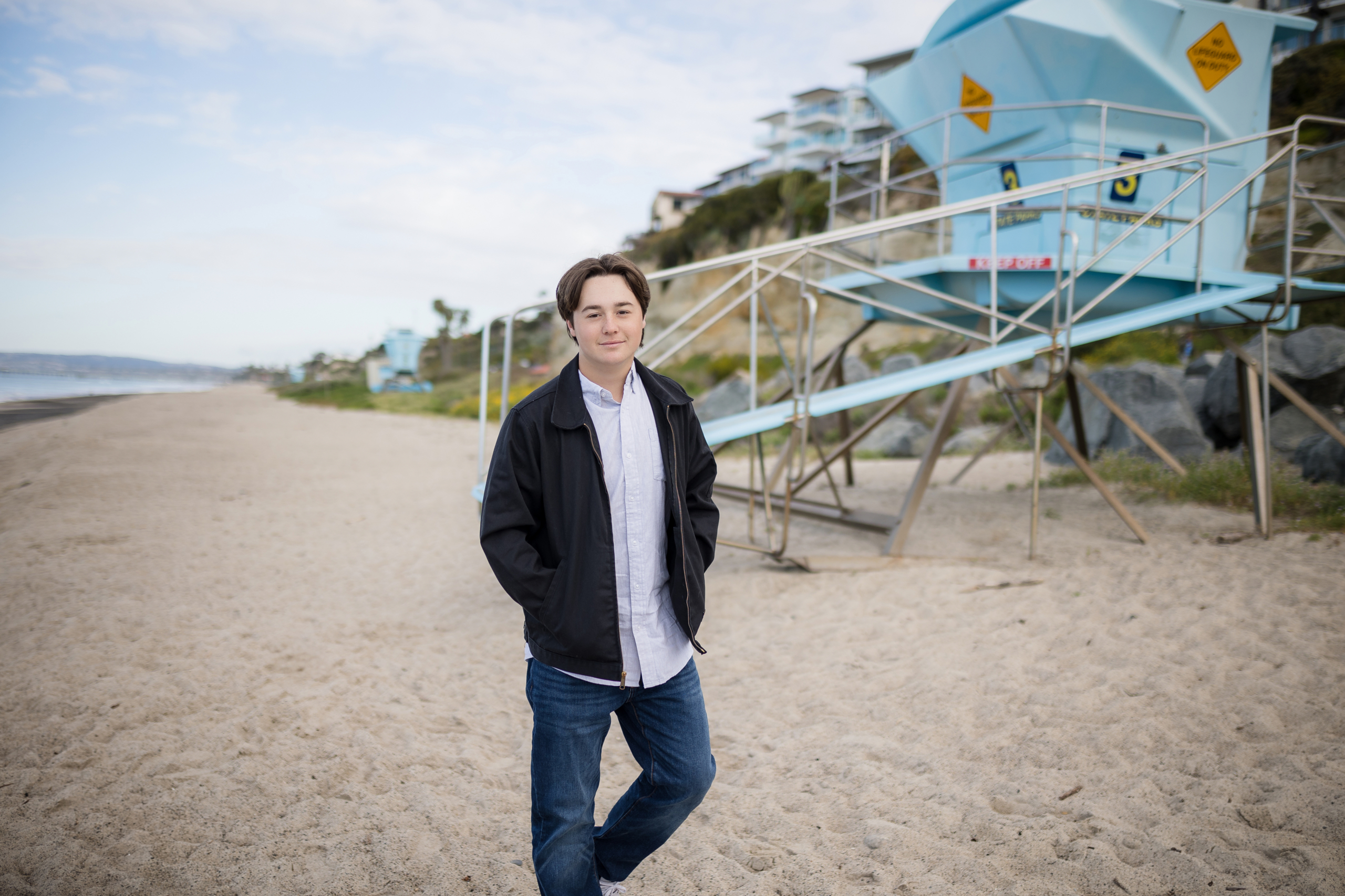 A high school senior leans casually against a lifeguard tower on the sand, dressed in a black jacket over a white button-up shirt. The ocean, cliffs, and beachfront homes add depth to the coastal setting.