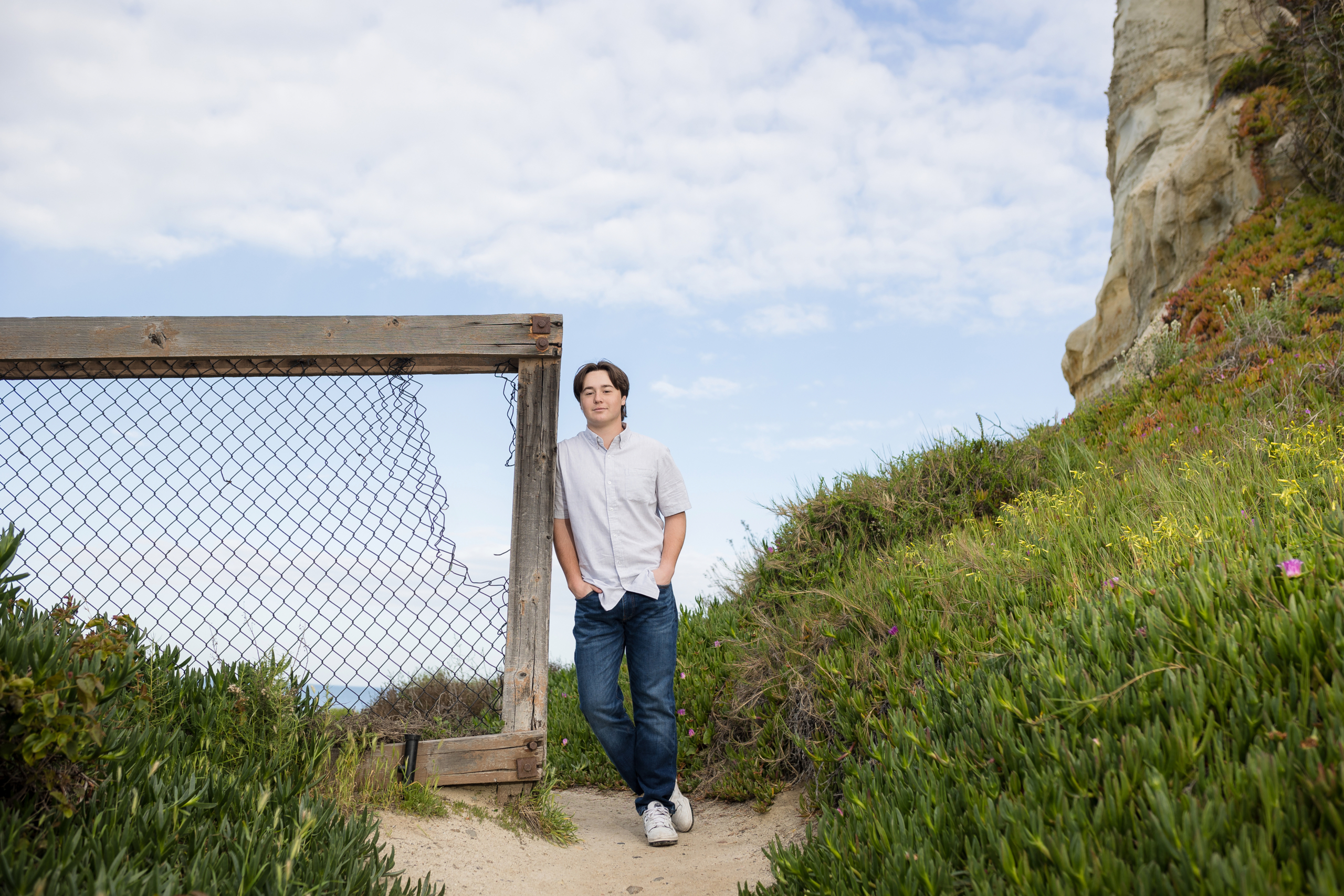 Senior boy leaning against a rustic wooden fence with a broken wire section, surrounded by lush green hills and wildflowers, with a towering cliffside and a bright blue sky overhead.