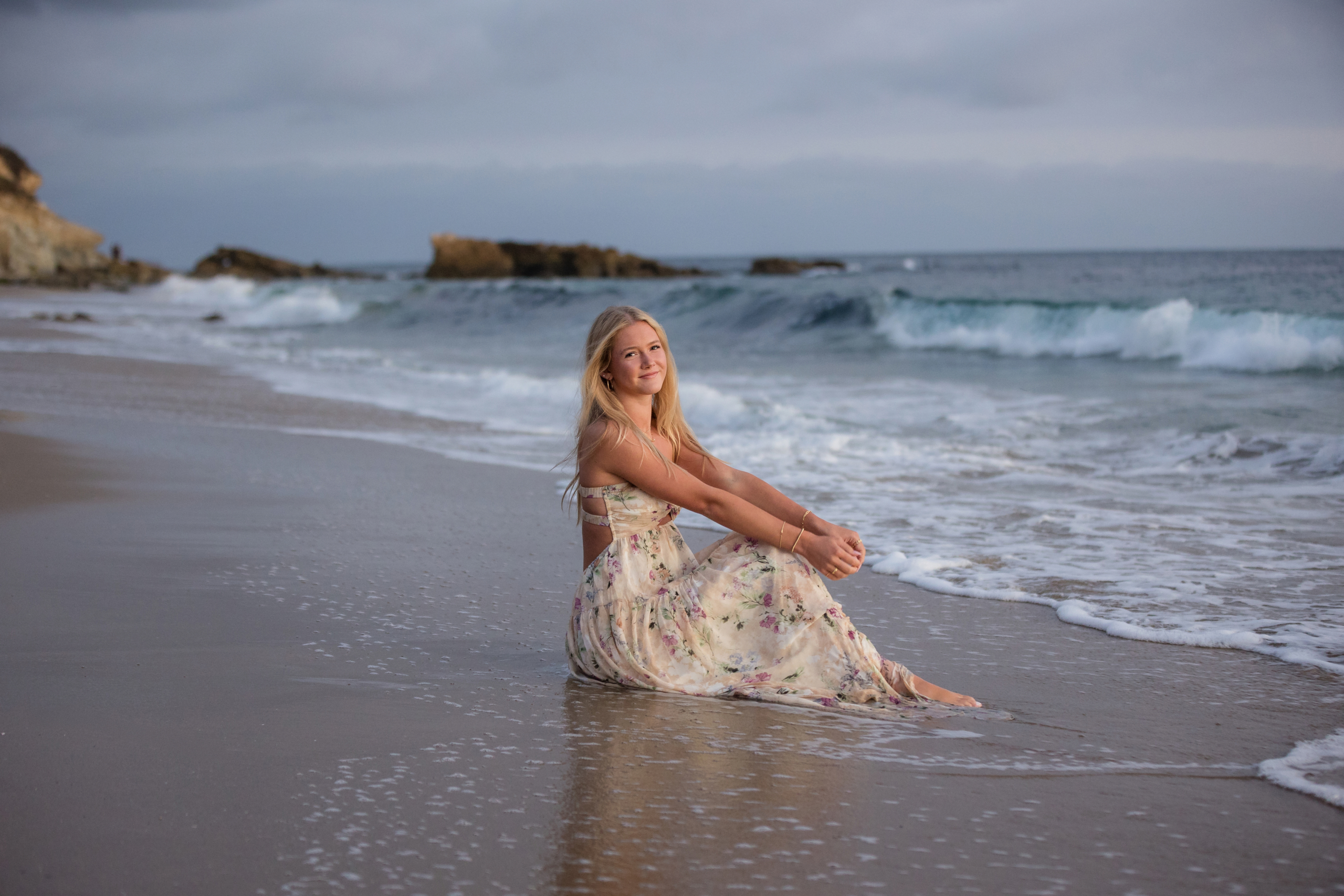 A young woman in a floral dress sits on the wet sand near the ocean, smiling gently as the waves touch her feet, framed by a moody sky.