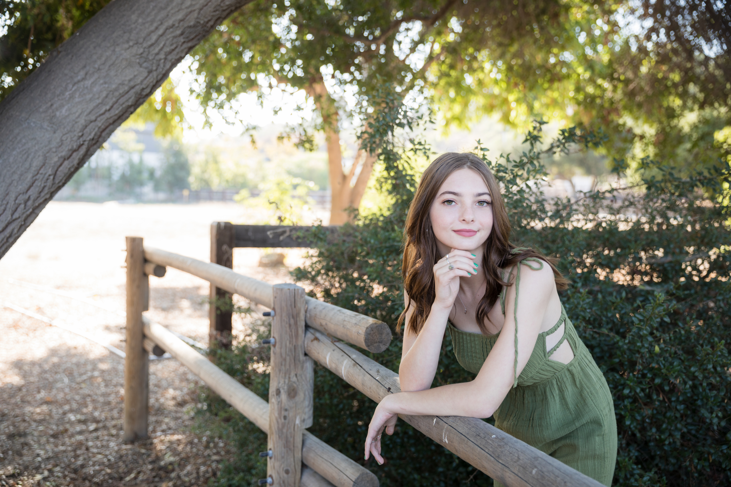 High school senior girl in a green dress leaning on a rustic wooden fence, surrounded by greenery with warm sunlight filtering through the trees. A soft, natural setting for senior portraits in Orange County