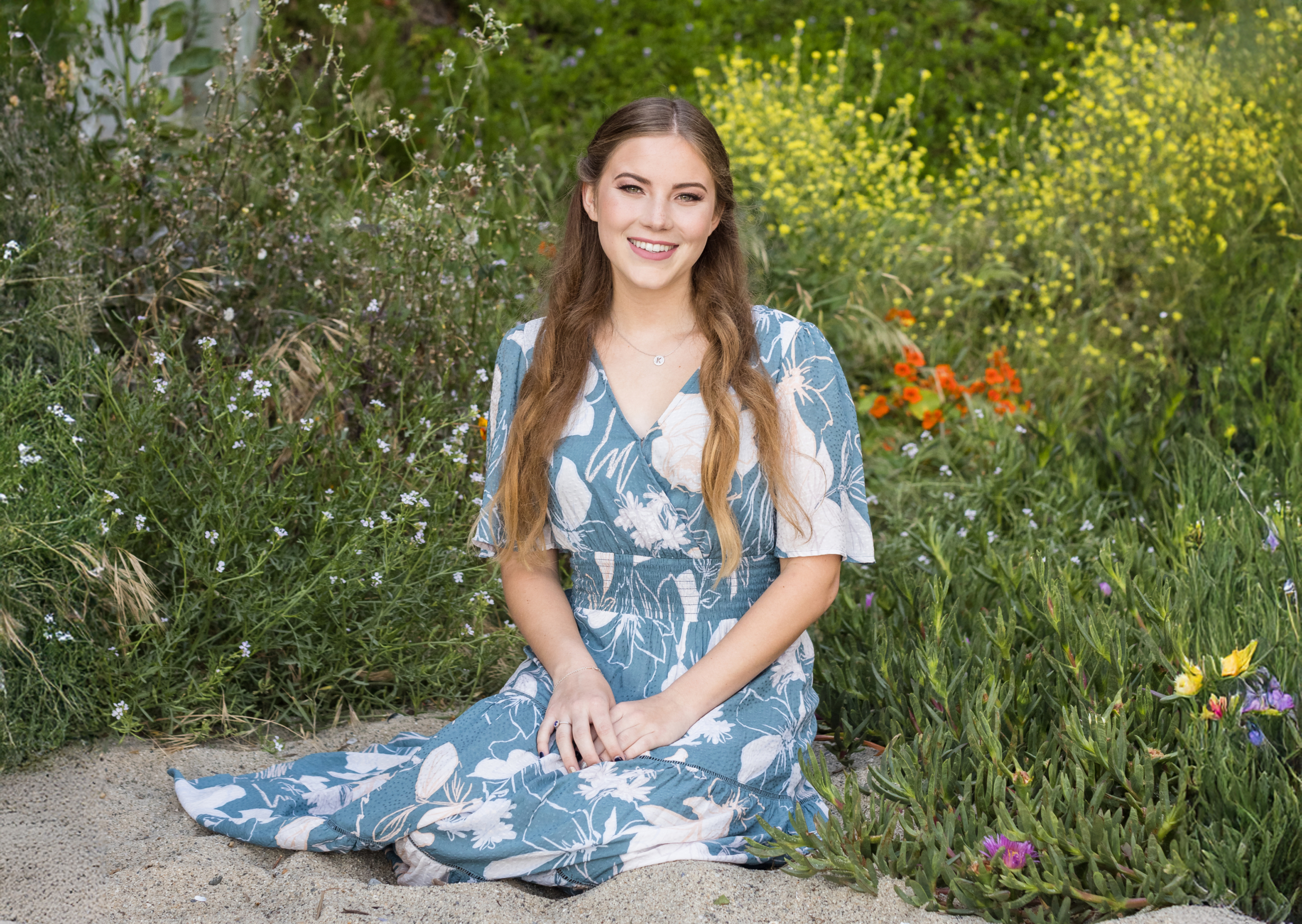 High school senior girl with long wavy hair wearing a blue floral dress, sitting on a sandy path surrounded by colorful wildflowers. A bright and cheerful outdoor senior portrait in Orange County.