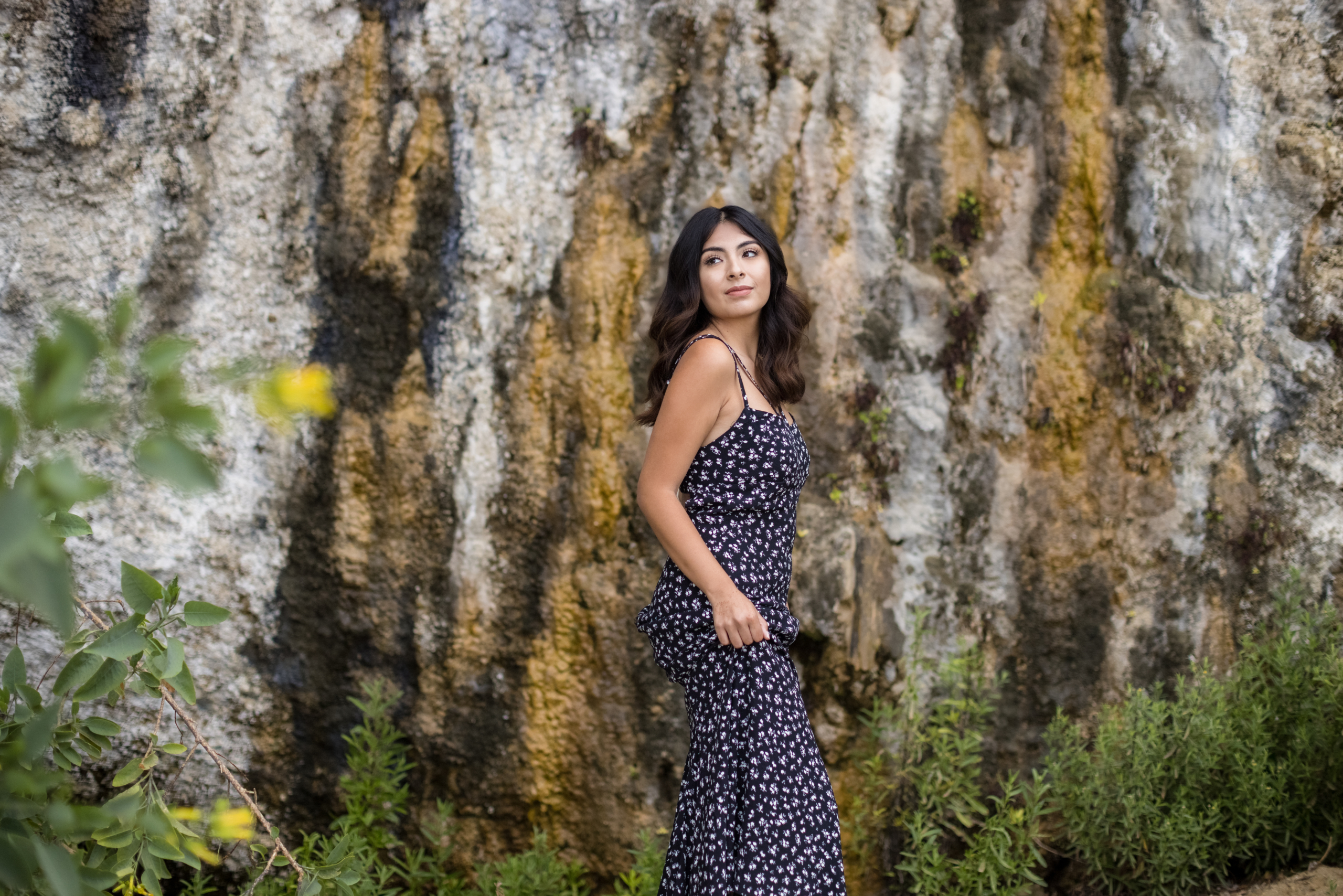 High school senior girl in a floral maxi dress standing against a rugged, textured rock wall, surrounded by greenery. A stylish and earthy setting for senior portraits in Orange County.