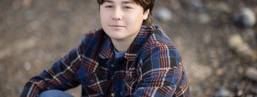 A senior boy stands confidently on a quiet beach, wearing a gray Emerson hoodie with his hands in his pockets. The soft waves roll onto the shore under a blue sky with wispy clouds, creating a peaceful backdrop.