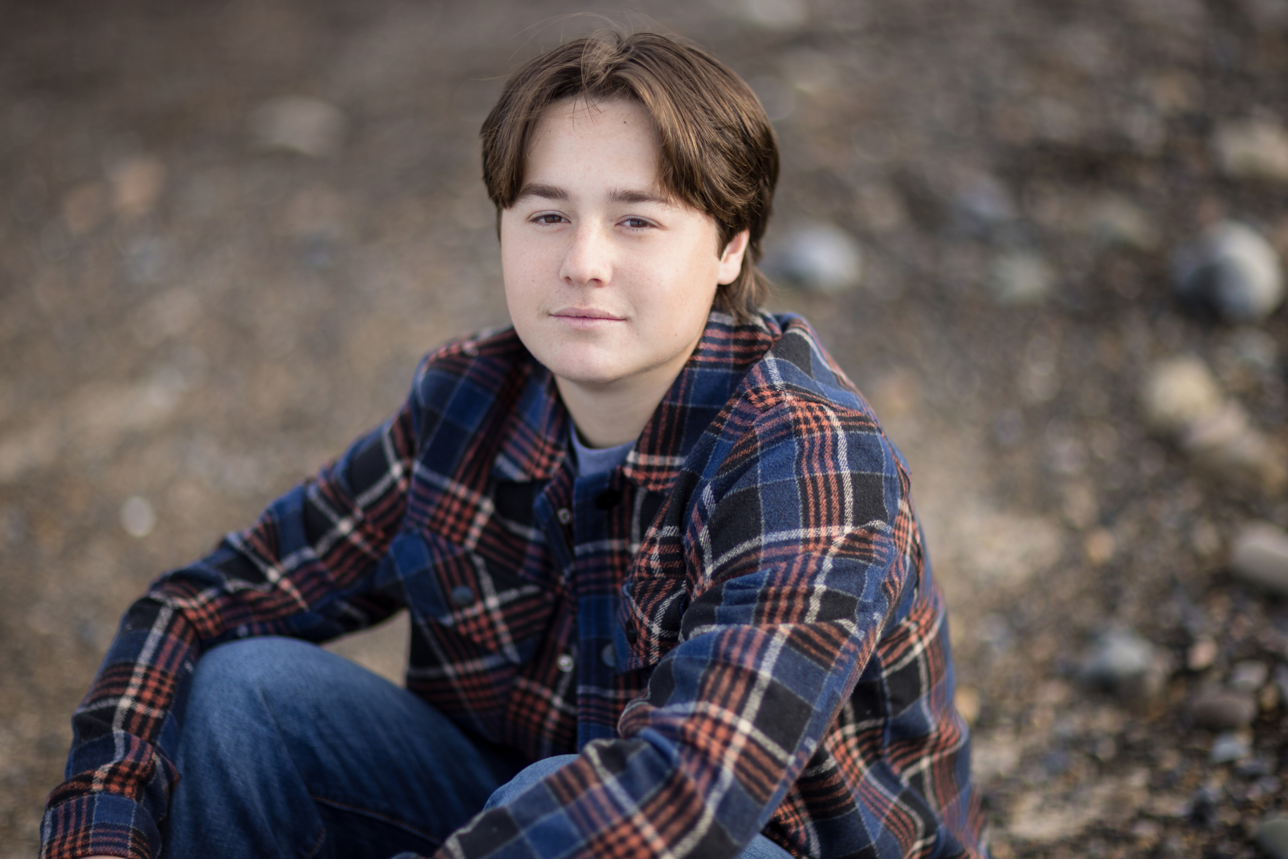 A senior boy stands confidently on a quiet beach, wearing a gray Emerson hoodie with his hands in his pockets. The soft waves roll onto the shore under a blue sky with wispy clouds, creating a peaceful backdrop.