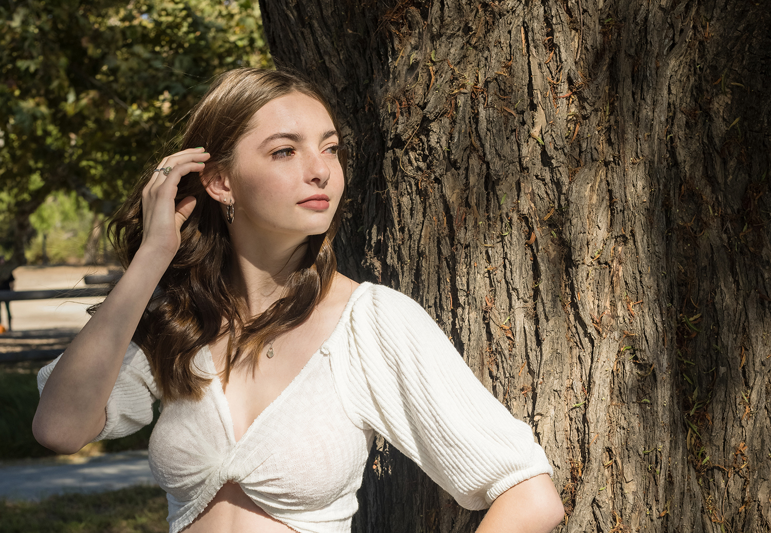 High school senior girl with wavy brown hair wearing a white cropped top, leaning against a textured tree trunk while gazing into the distance. Sunlight and shadows create a warm, natural senior portrait in Orange County.