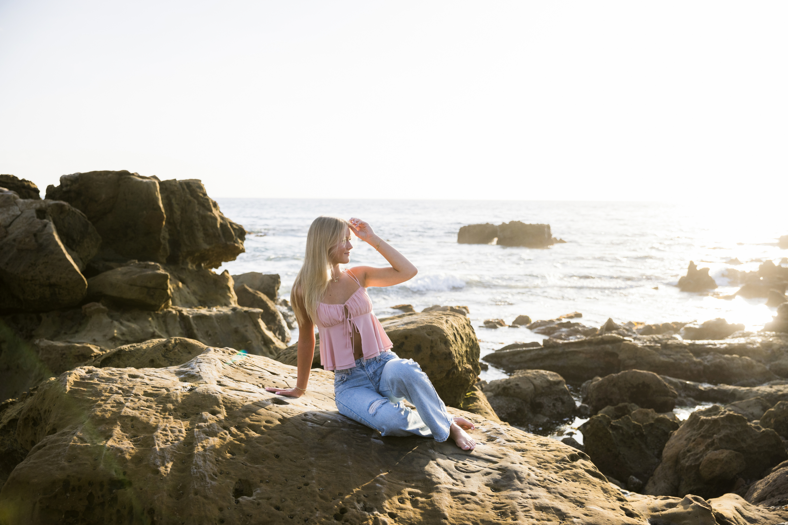 A senior girl in a pink top and jeans sits on a large rock near the shoreline, shielding her eyes from the bright golden hour sun while gazing into the distance.