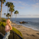 A high school senior leans against a coastal railing, overlooking a scenic beach framed by palm trees, with the ocean stretching into the horizon behind her.