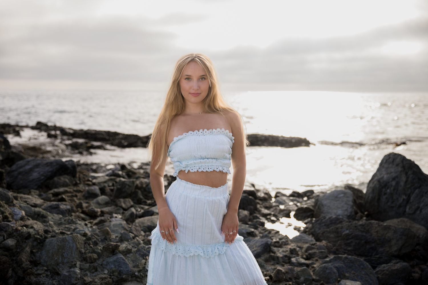 Blonde high school senior standing on a rocky shoreline at sunset, wearing a white eyelet two-piece outfit. The ocean and glowing sky create a dreamy, golden-hour backdrop for this Laguna Beach senior portrait.