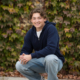 Smiling high school senior crouching in front of a lush ivy-covered wall, wearing a navy hoodie and light-wash jeans. A natural and relaxed pose for a professional senior portrait.