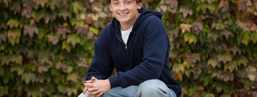 Smiling high school senior crouching in front of a lush ivy-covered wall, wearing a navy hoodie and light-wash jeans. A natural and relaxed pose for a professional senior portrait.